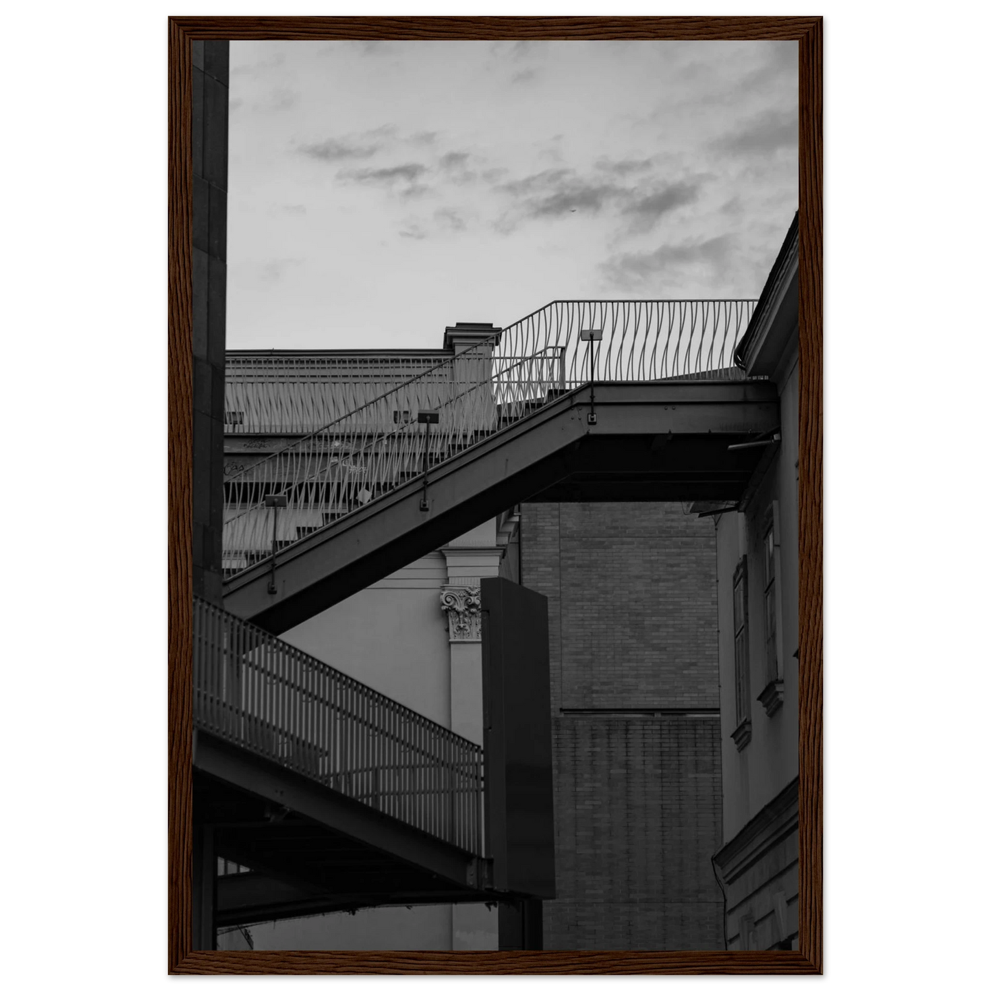 Black and white photo on a wooden frame wall art of an outdoor staircase connecting two buildings under a dramatic cloudy sky