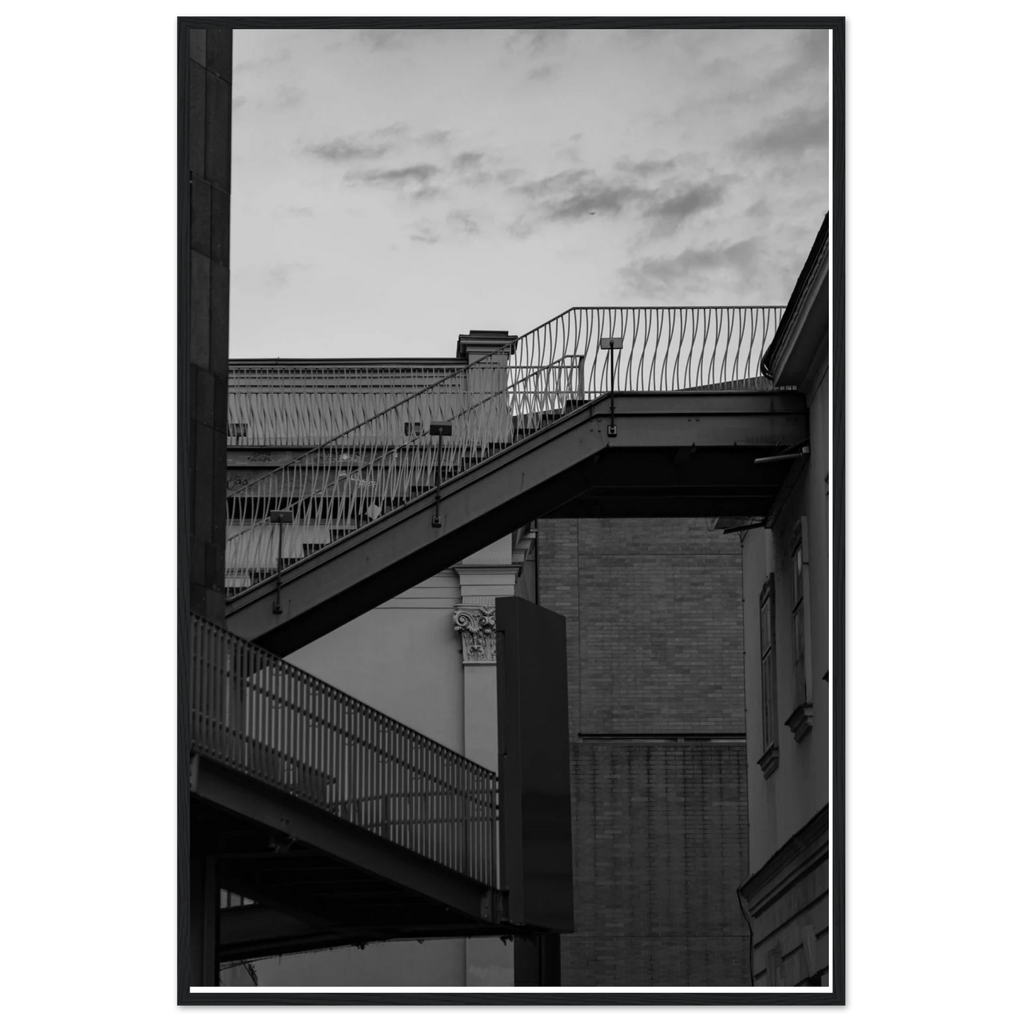 Black and white photo on a wooden frame wall art of an outdoor staircase connecting two buildings under a dramatic cloudy sky
