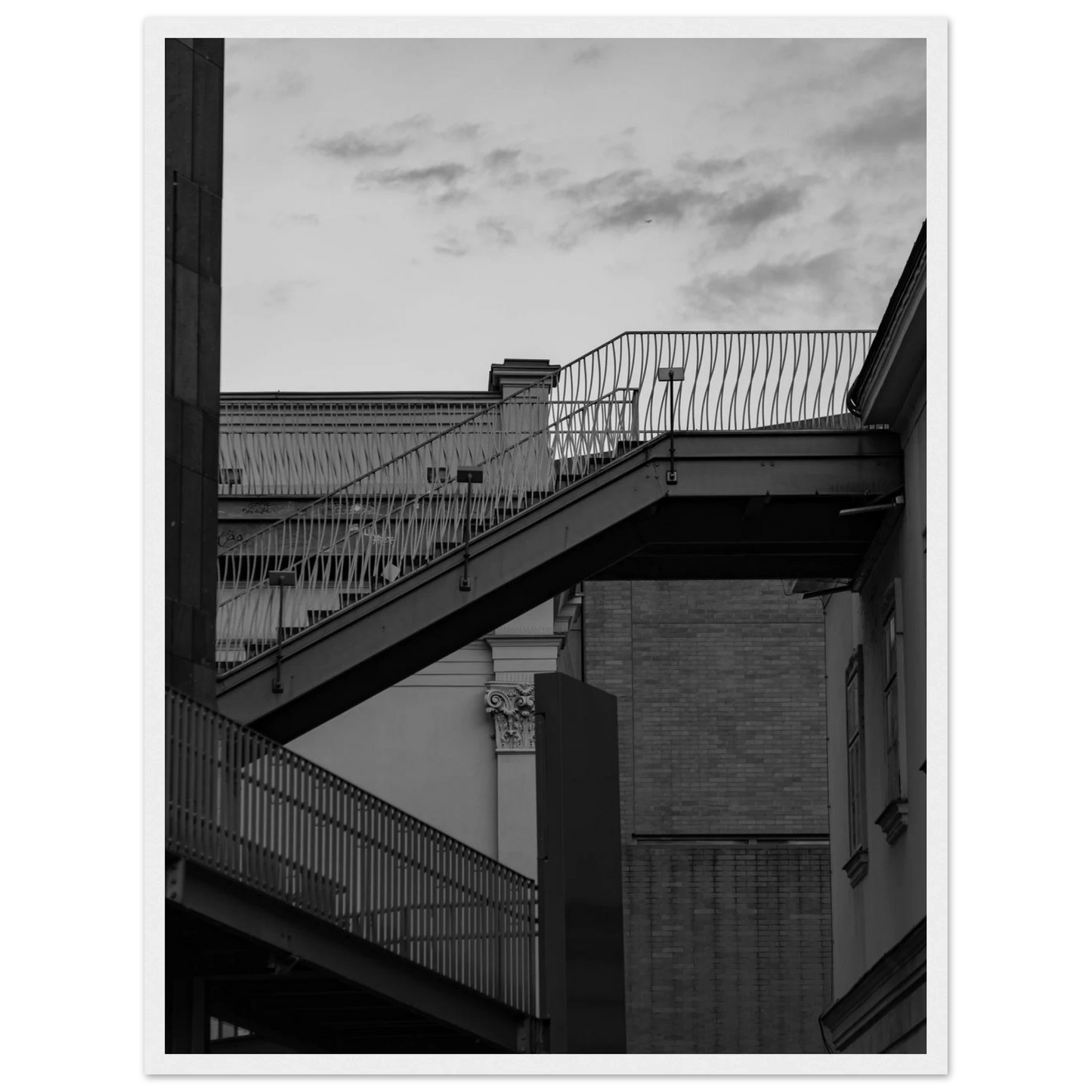 Black and white photo on a wooden frame wall art of an outdoor staircase connecting two buildings under a dramatic cloudy sky