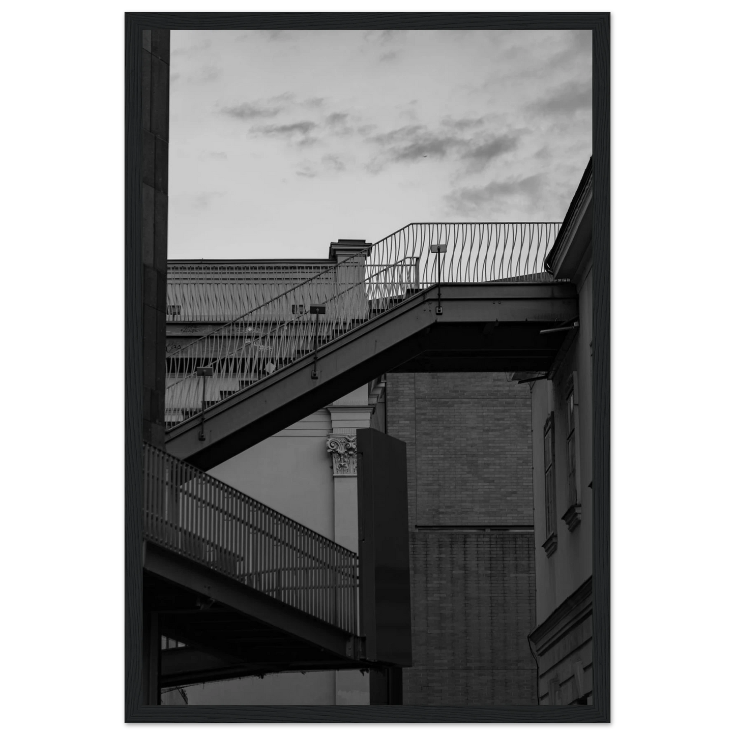 Black and white photo on a wooden frame wall art of an outdoor staircase connecting two buildings under a dramatic cloudy sky
