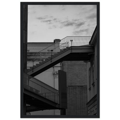 Black and white photo on a wooden frame wall art of an outdoor staircase connecting two buildings under a dramatic cloudy sky