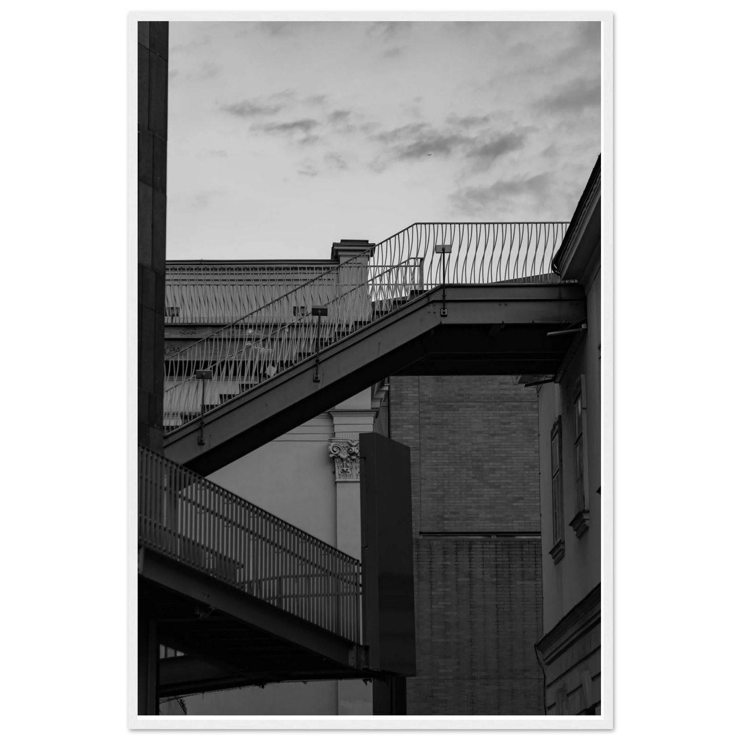 Black and white photo on a wooden frame wall art of an outdoor staircase connecting two buildings under a dramatic cloudy sky