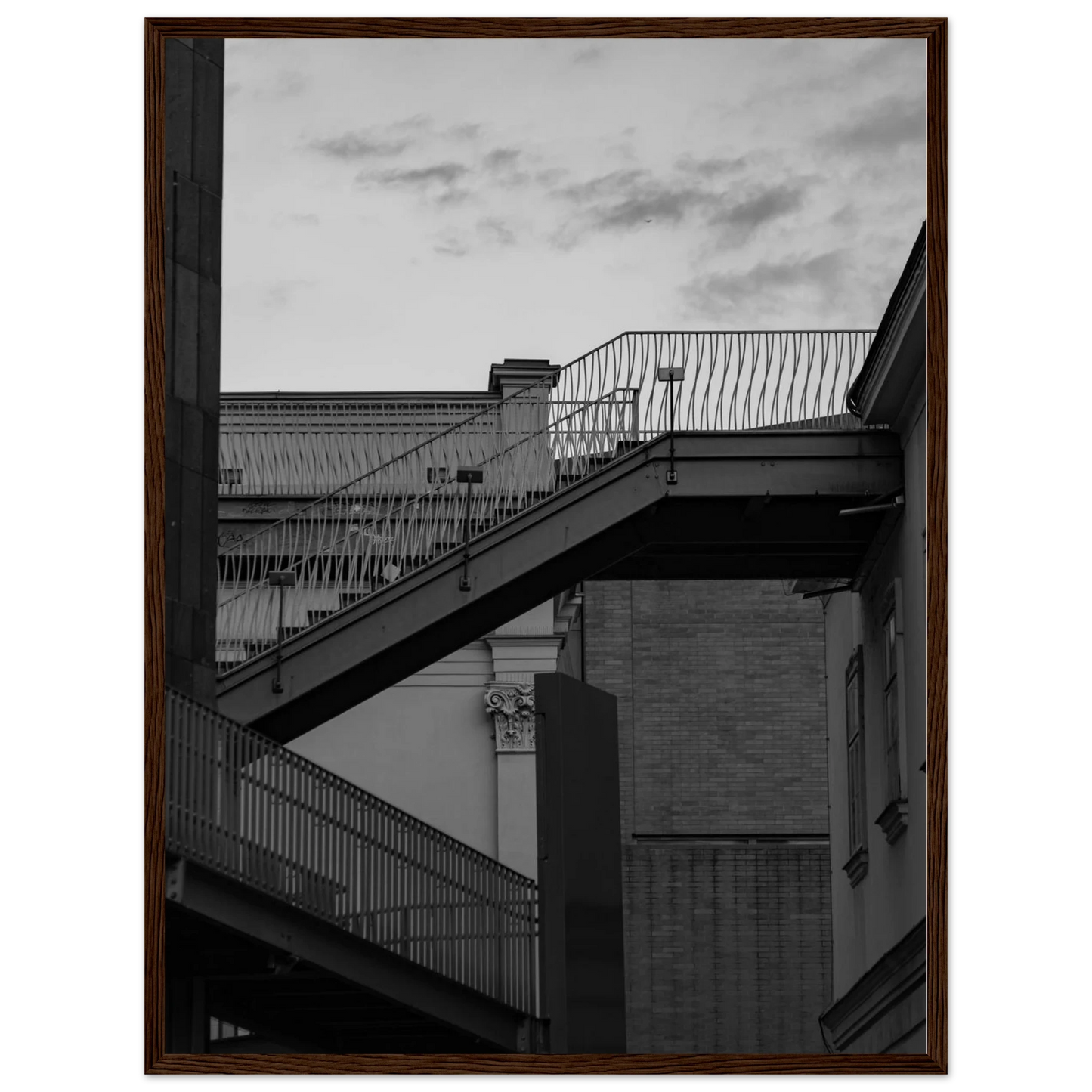 Black and white photo on a wooden frame wall art of an outdoor staircase connecting two buildings under a dramatic cloudy sky