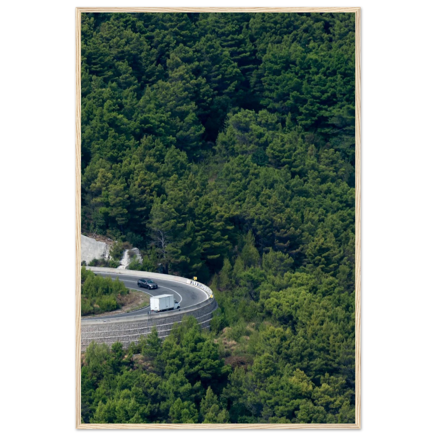 Aerial view of a winding road curve with cars surrounded by lush green forest landscape on wooden framed wall art