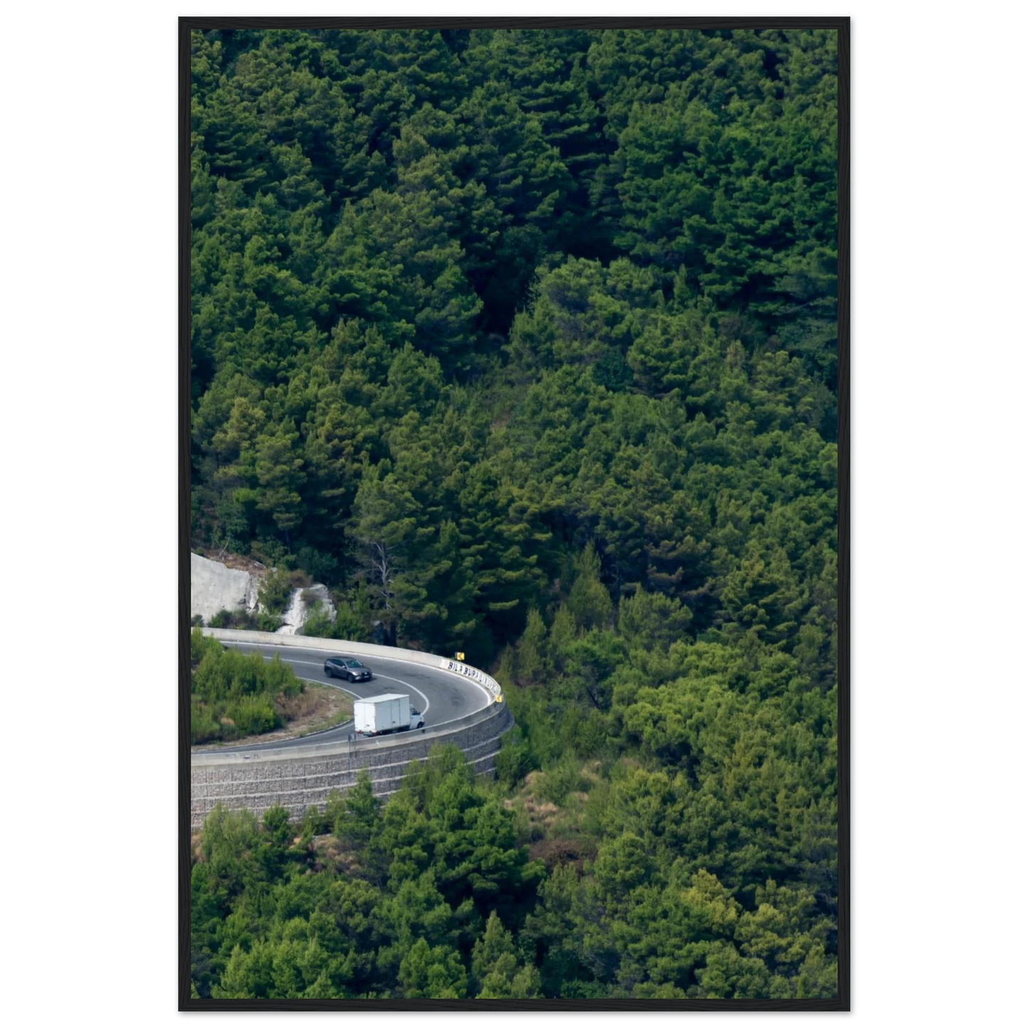 Aerial view of a winding road curve with cars surrounded by lush green forest landscape on wooden framed wall art