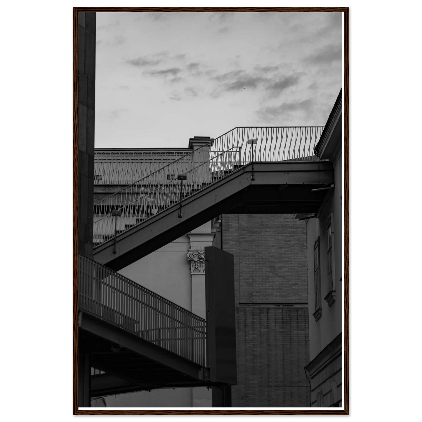 Black and white photo on a wooden frame wall art of an outdoor staircase connecting two buildings under a dramatic cloudy sky