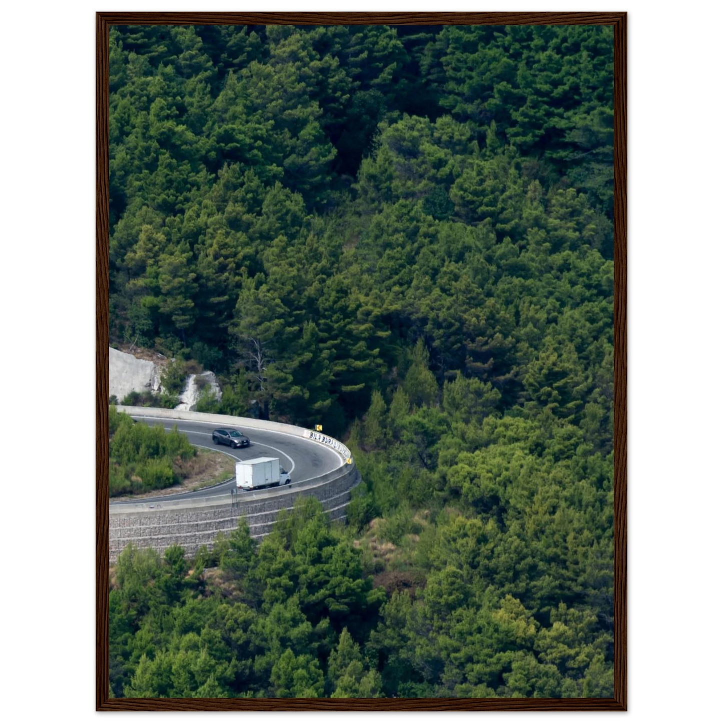 Aerial view of a winding road curve with cars surrounded by lush green forest landscape on wooden framed wall art
