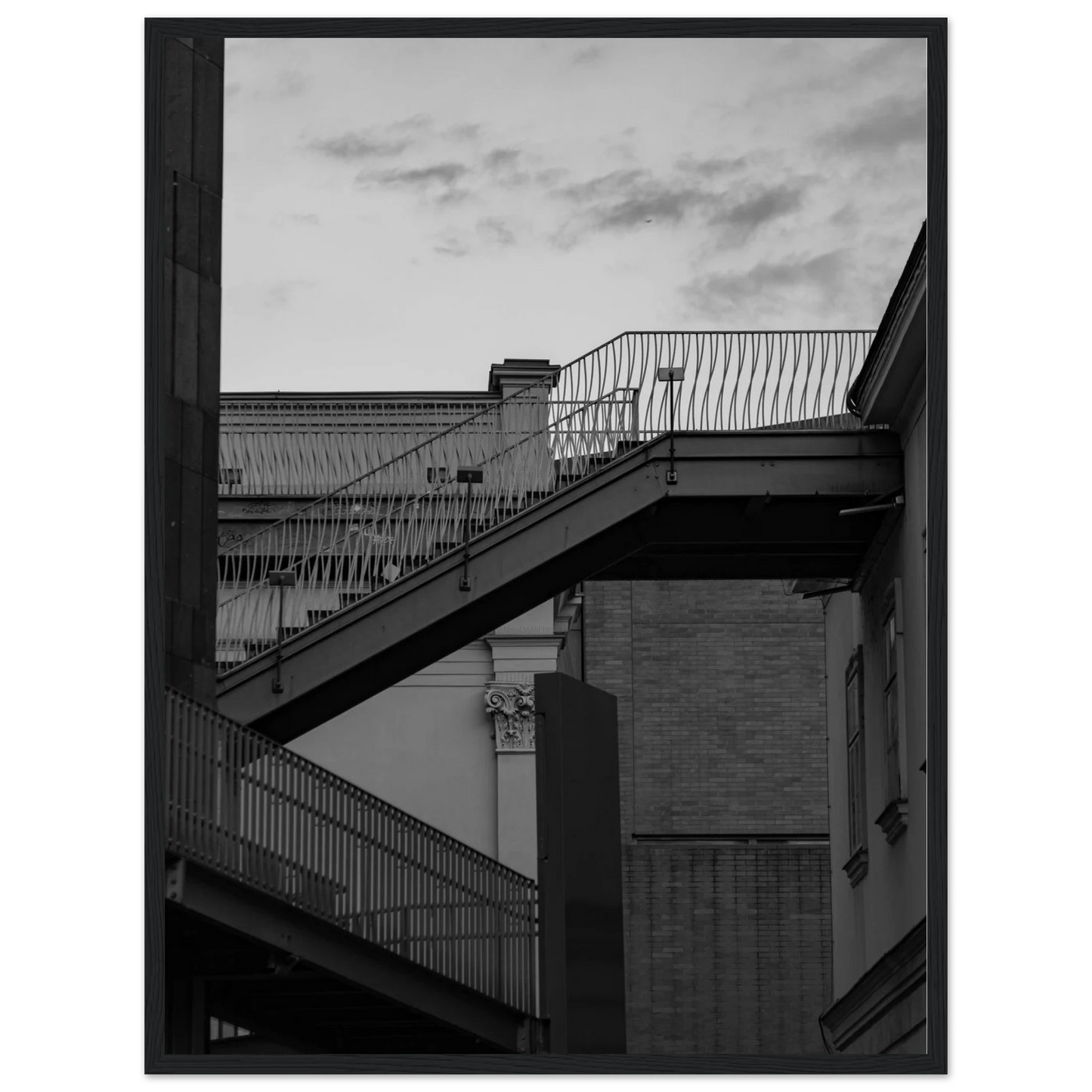 Black and white photo on a wooden frame wall art of an outdoor staircase connecting two buildings under a dramatic cloudy sky