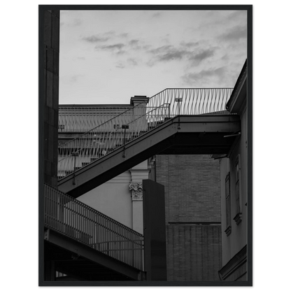 Black and white photo on a wooden frame wall art of an outdoor staircase connecting two buildings under a dramatic cloudy sky