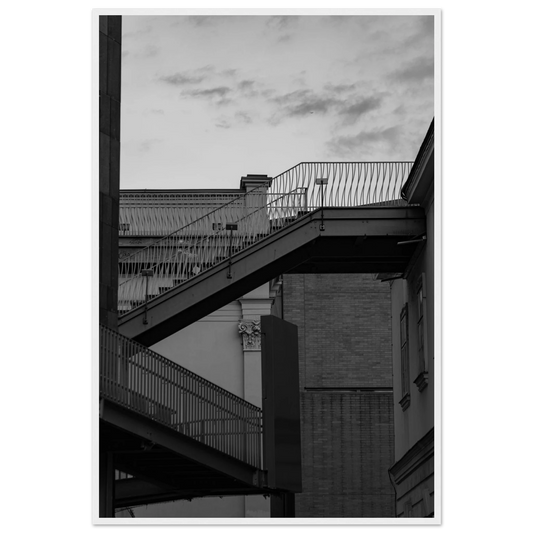 Black and white photo on a wooden frame wall art of an outdoor staircase connecting two buildings under a dramatic cloudy sky
